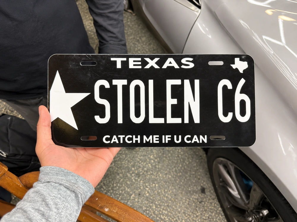 The STOLEN C6 Texas-style custom plate held beside a silver car.