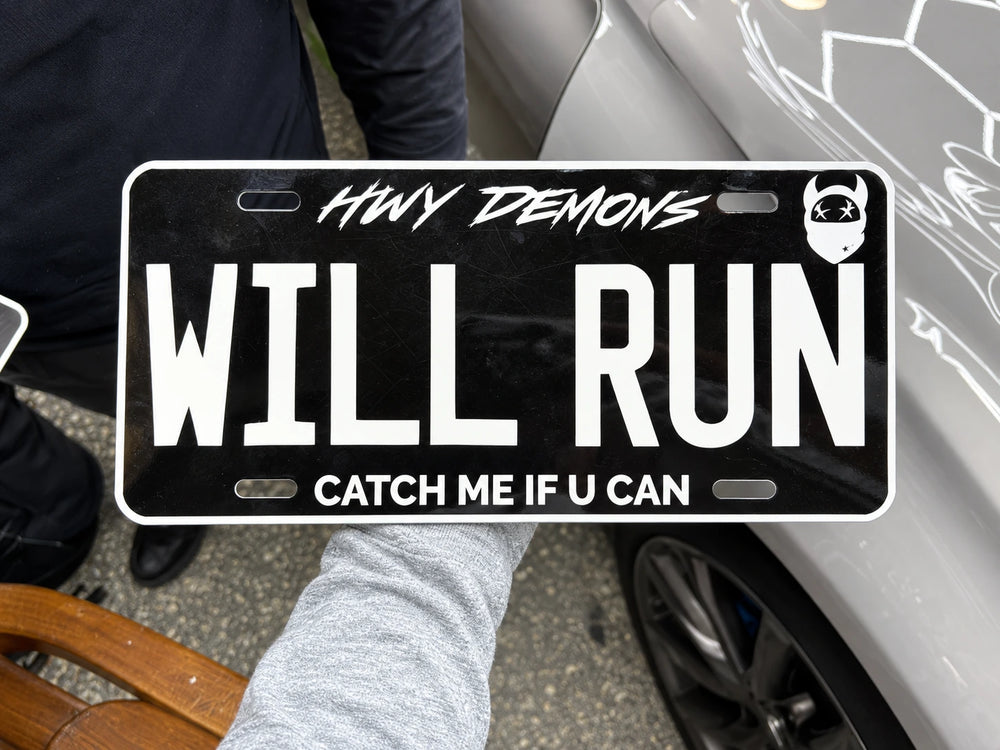 The HWY DEMONS WILL RUN custom plate held beside a silver car.