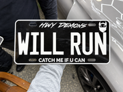 The HWY DEMONS WILL RUN custom plate held beside a silver car.